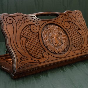 Könnte beinhalten: Ein Backgammon-Spielbrett aus Holz mit aufwendigen Schnitzereien. Das Brett zeigt ein zentrales Löwenkopf-Design, umgeben von verzierten Mustern und einem Griff zum Tragen. Das dunkelbraune Holz hat eine polierte Oberfläche, die die detaillierte Handwerkskunst hervorhebt.