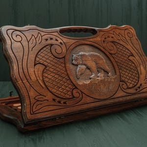 May include: A wooden backgammon set with a carved bear design. The set is made of dark brown wood and features intricate carvings and a handle for carrying. The bear is in an oval frame.