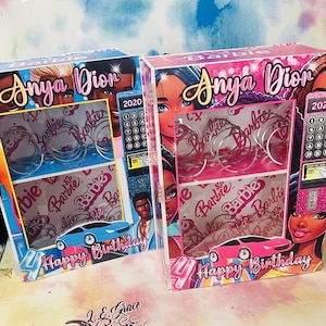Könnte beinhalten: Zwei personalisierte Geschenkboxen in Pink und Blau mit dem Namen "Anya Dion" und dem Jahr "2020" darauf. Die Boxen haben ein Barbie-Thema mit Bildern von Barbie-Puppen und dem Wort "Barbie", das auf den Boxen wiederholt wird. Die Boxen haben ein Autobild unten und die Worte "Happy Birthday" auf der Vorderseite.