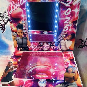 May include: A pink and white vanity mirror with a lighted frame. The mirror has a pink background with a large pink lip design. The vanity mirror has a clear glass top and a pink base. The mirror has a sign that says "Laila & Grace Designs" and "Happy Easter 2025".