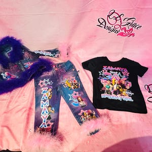 May include: A denim jacket and jeans set with a black t-shirt. The jacket and jeans are decorated with cartoon characters, stars, and the name "ZAMAYA". The jacket has purple fur trim, and the jeans have pink fur trim at the ankles. The black t-shirt says "ZAMAYA 3rd BIRTHDAY GIRL".