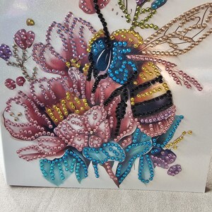 Op de afbeelding: Een decoratief kunstwerk met een bij en bloemen, versierd met kleurrijke strass-steentjes. De bij heeft een blauwe kop, een geel en zwart gestreept lichaam en gedetailleerde vleugels. De bloemen zijn roze, blauw en paars, wat een levendig ontwerp cre&euml;ert.