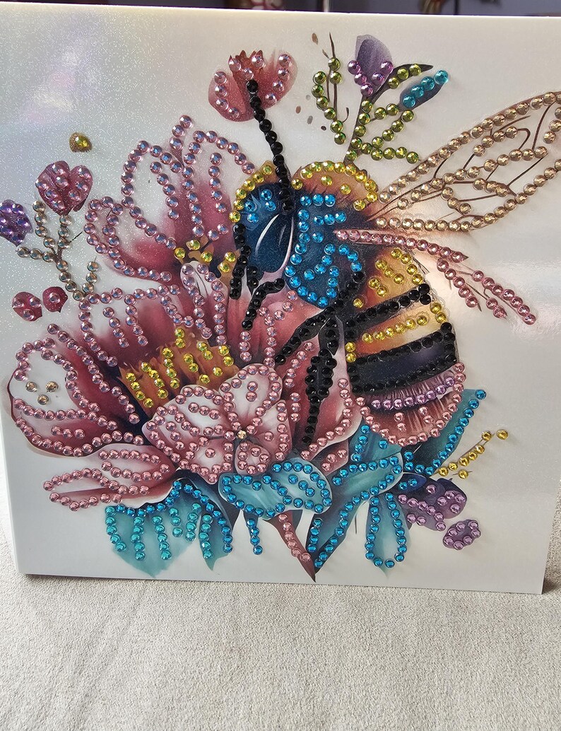 Op de afbeelding: Een decoratief kunstwerk met een bij en bloemen, versierd met kleurrijke strass-steentjes. De bij heeft een blauwe kop, een geel en zwart gestreept lichaam en gedetailleerde vleugels. De bloemen zijn roze, blauw en paars, wat een levendig ontwerp cre&euml;ert.