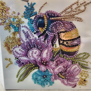 Op de afbeelding: Een decoratief kunstwerk met een bij en een bloemenontwerp. De bij is versierd met paarse, zwarte en gouden elementen, en de bloemen zijn in tinten paars, blauw en goud. Het geheel is geplaatst tegen een witte achtergrond, wat een opvallend contrast cre&euml;ert.