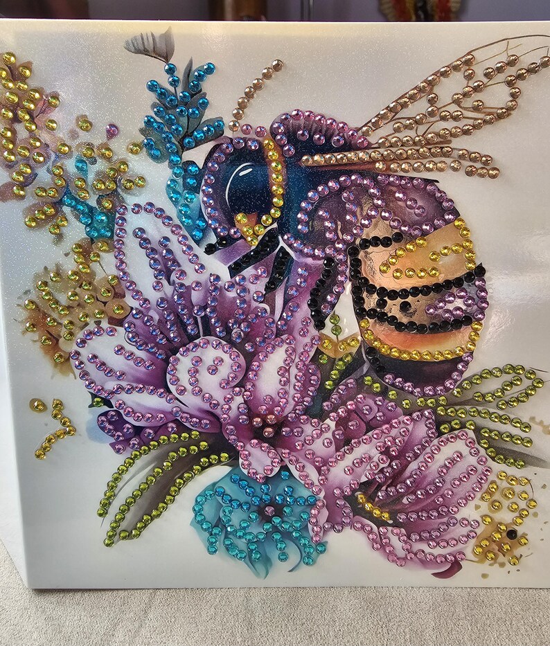 Op de afbeelding: Een decoratief kunstwerk met een bij en een bloemenontwerp. De bij is versierd met paarse, zwarte en gouden elementen, en de bloemen zijn in tinten paars, blauw en goud. Het geheel is geplaatst tegen een witte achtergrond, wat een opvallend contrast cre&euml;ert.