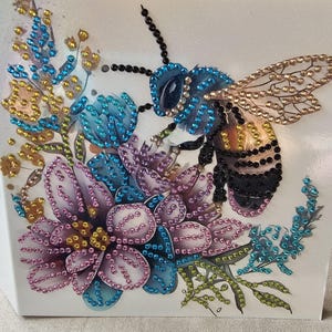 Op de afbeelding: Decoratieve afbeelding met een gedetailleerde bij en bloemen, versierd met sprankelende strass-steentjes. De bij is blauw en zwart, met goudkleurige vleugels, in de buurt van paarse, blauwe en gele bloemen. Het kunstwerk staat op een witte achtergrond.