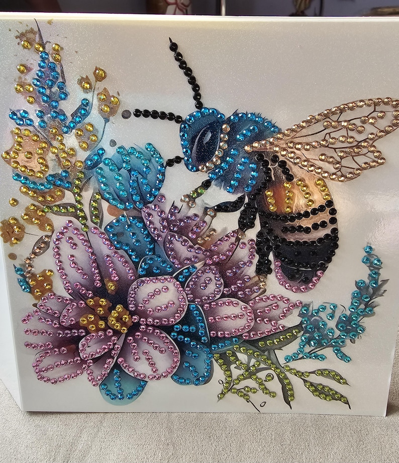 Op de afbeelding: Decoratieve afbeelding met een gedetailleerde bij en bloemen, versierd met sprankelende strass-steentjes. De bij is blauw en zwart, met goudkleurige vleugels, in de buurt van paarse, blauwe en gele bloemen. Het kunstwerk staat op een witte achtergrond.