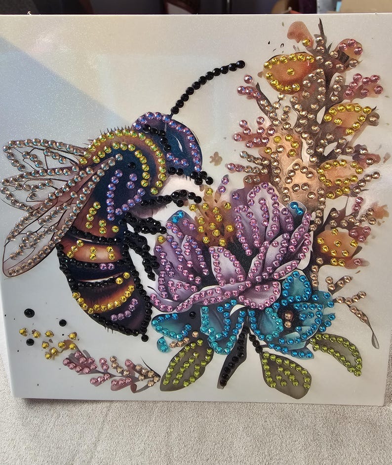 Op de afbeelding: Een decoratief kunstwerk met een grote bij en bloemen, versierd met kleurrijke strass-steentjes. De bij heeft een zwart en geel lichaam met blauwe en paarse vleugels. De bloemen zijn paars, blauw en oranje, met groene bladeren.