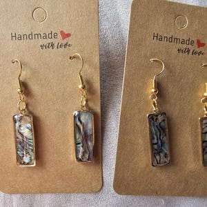 Può includere: Due paia di orecchini fatti a mano con ganci color oro e pendenti rettangolari. I pendenti presentano conchiglia di abalone iridescente incastonata in metallo color oro. Gli orecchini sono esposti su cartoncini marroni con la scritta "Handmade with love."