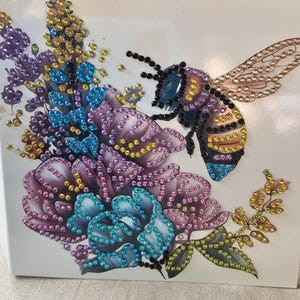 Op de afbeelding: Een decoratief kunstwerk met een grote bij en bloemen, versierd met kleurrijke strass-steentjes. De bij heeft zwarte, gele en blauwe accenten, terwijl de bloemen tinten paars, blauw en geel vertonen. Het kunstwerk staat op een witte achtergrond.