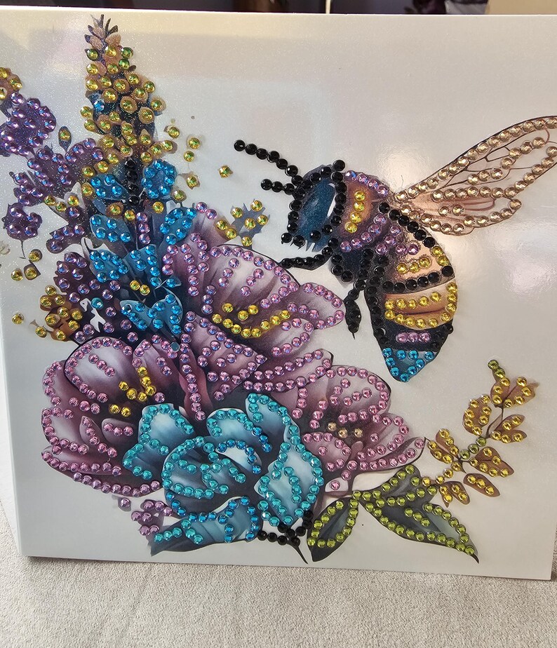 Op de afbeelding: Een decoratief kunstwerk met een grote bij en bloemen, versierd met kleurrijke strass-steentjes. De bij heeft zwarte, gele en blauwe accenten, terwijl de bloemen tinten paars, blauw en geel vertonen. Het kunstwerk staat op een witte achtergrond.