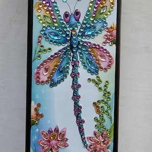 May include: A black phone case featuring a colourful dragonfly design made of rhinestones. The dragonfly has blue, green, pink and yellow rhinestones. The background is a light blue with flowers made of rhinestones.