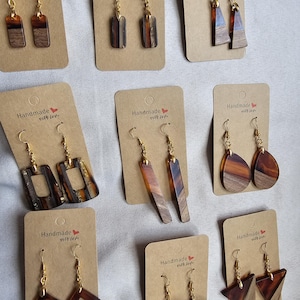Puede incluir: Una colección de pendientes hechos a mano exhibidos en tarjetas marrones. Los pendientes presentan varias formas y diseños geométricos, incorporando madera y resina en tonos marrón, ámbar y dorado. Cada tarjeta tiene el texto "Handmade with love".