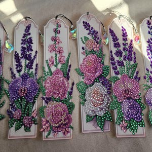 May include: Six colourful bookmarks featuring different floral designs. Each bookmark is decorated with rhinestones and has a small crystal charm hanging from the top.