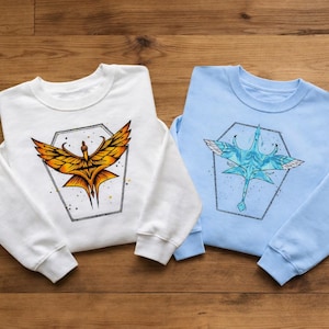 May include: Two crew neck sweatshirts, one white and one light blue. Each features a stylized bird design within a hexagonal frame. The white sweatshirt has a gold and orange bird, while the blue one has a blue and white bird.