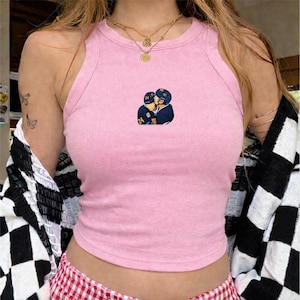 May include: A light pink tank top with a small embroidered design of two people in hockey uniforms embracing. The top is sleeveless and has a round neckline. A black and white checkered cardigan is draped over the shoulders.
