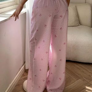 May include: Wide-leg lounge pants featuring a light pink and white vertical striped pattern with small, scattered pink bow icons. The pants have an elasticized waistband and a textured, seersucker-like fabric finish throughout the garment.