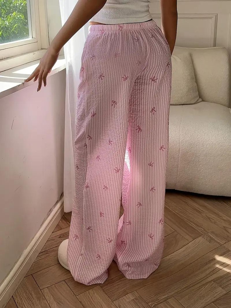 May include: Wide-leg lounge pants featuring a light pink and white vertical striped pattern with small, scattered pink bow icons. The pants have an elasticized waistband and a textured, seersucker-like fabric finish throughout the garment.