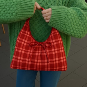 May include: A red plaid handbag with a bow detail. The bag is held by a person wearing a green sweater and blue jeans. The bag's pattern is a mix of red shades, and the bow adds a decorative touch.