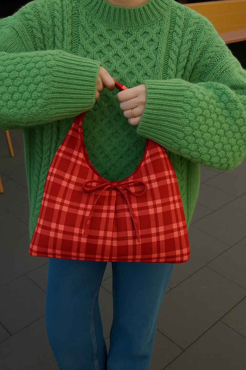 May include: A red plaid handbag with a bow detail. The bag is held by a person wearing a green sweater and blue jeans. The bag's pattern is a mix of red shades, and the bow adds a decorative touch.