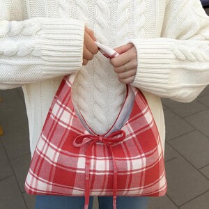 May include: A red and white plaid shoulder bag with a bow closure. The bag is held by a person wearing a white cable-knit sweater and blue jeans. The bag's design is a classic gingham pattern.