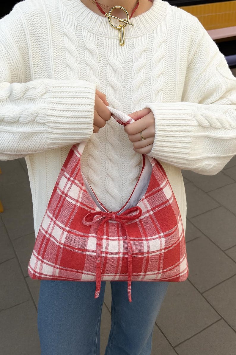 May include: A red and white plaid shoulder bag with a bow closure. The bag is held by a person wearing a white cable-knit sweater and blue jeans. The bag's design is a classic gingham pattern.