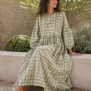 Pode incluir: Um vestido de manga comprida at&eacute; ao ch&atilde;o com um padr&atilde;o de xadrez vichy em verde s&aacute;lvia e branco. A pe&ccedil;a apresenta um decote redondo, uma cintura franzida e mangas volumosas com punhos el&aacute;sticos, criando uma silhueta solta e fluida.