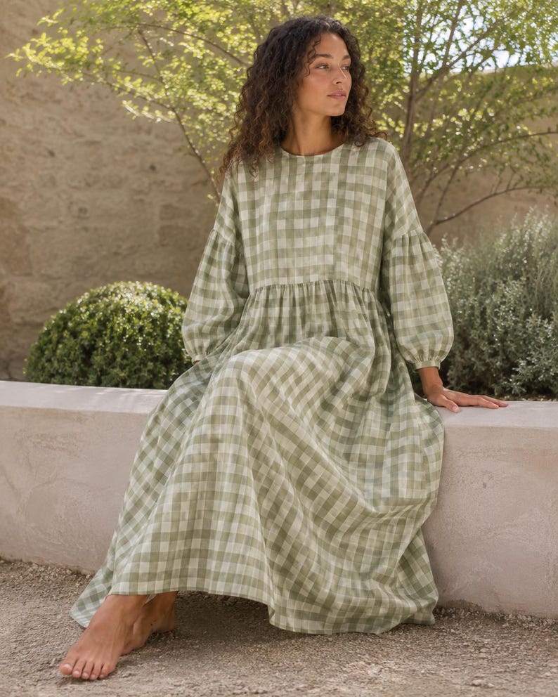 Pode incluir: Um vestido de manga comprida at&eacute; ao ch&atilde;o com um padr&atilde;o de xadrez vichy em verde s&aacute;lvia e branco. A pe&ccedil;a apresenta um decote redondo, uma cintura franzida e mangas volumosas com punhos el&aacute;sticos, criando uma silhueta solta e fluida.