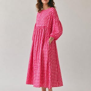 Pode incluir: Um vestido midi de manga comprida com um vibrante padr&atilde;o de xadrez vichy em rosa choque e branco. A pe&ccedil;a apresenta um decote redondo, uma cintura franzida e mangas volumosas com punhos el&aacute;sticos, criando uma silhueta solta e fluida.