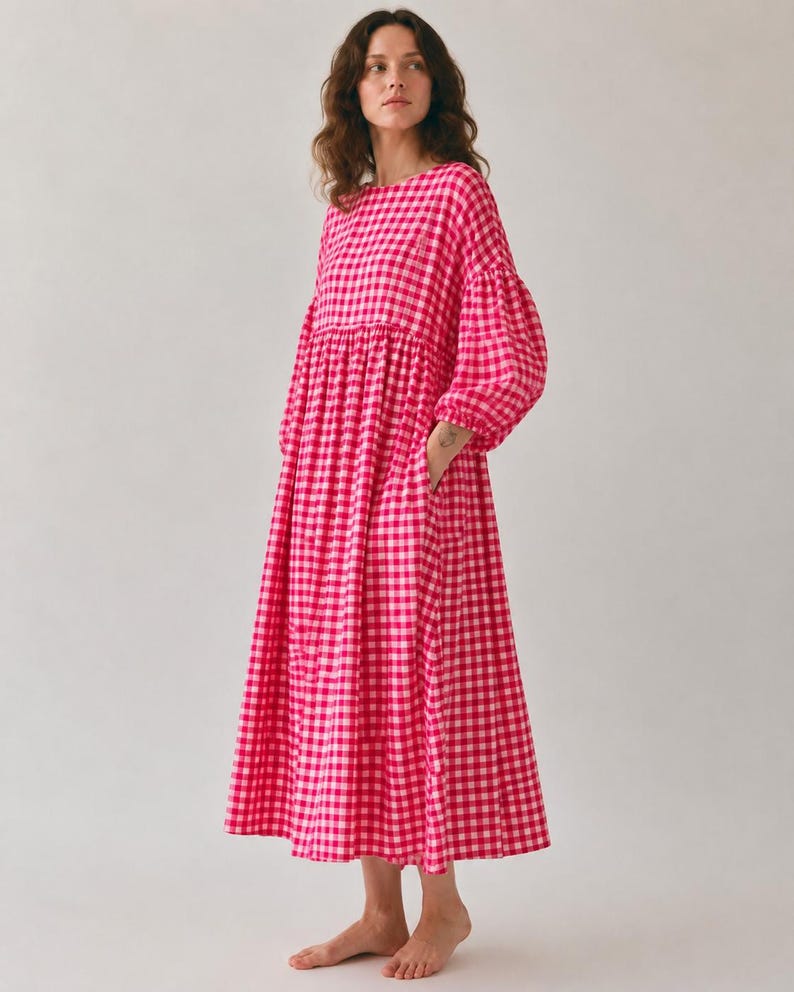 Pode incluir: Um vestido midi de manga comprida com um vibrante padr&atilde;o de xadrez vichy em rosa choque e branco. A pe&ccedil;a apresenta um decote redondo, uma cintura franzida e mangas volumosas com punhos el&aacute;sticos, criando uma silhueta solta e fluida.