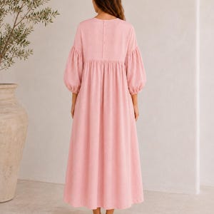 Pode incluir: Um vestido comprido rosa claro com decote redondo e uma fila vertical de bot&otilde;es na parte superior das costas. O vestido apresenta mangas bufantes volumosas at&eacute; ao cotovelo com punhos franzidos e uma saia larga e fluida.