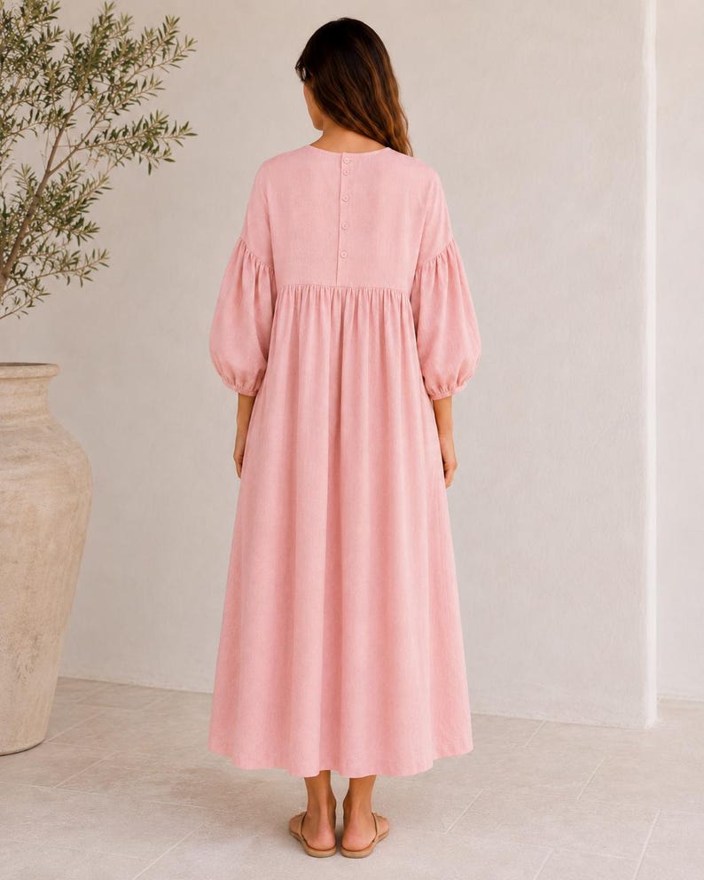 Pode incluir: Um vestido comprido rosa claro com decote redondo e uma fila vertical de bot&otilde;es na parte superior das costas. O vestido apresenta mangas bufantes volumosas at&eacute; ao cotovelo com punhos franzidos e uma saia larga e fluida.