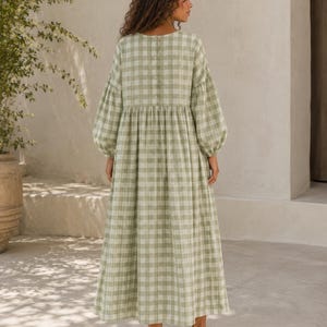 Pode incluir: Um vestido comprido at&eacute; aos p&eacute;s com um padr&atilde;o de xadrez vichy em verde s&aacute;lvia e branco. A pe&ccedil;a apresenta mangas compridas e volumosas com punhos franzidos, um decote redondo e uma fila vertical de bot&otilde;es no centro das costas.