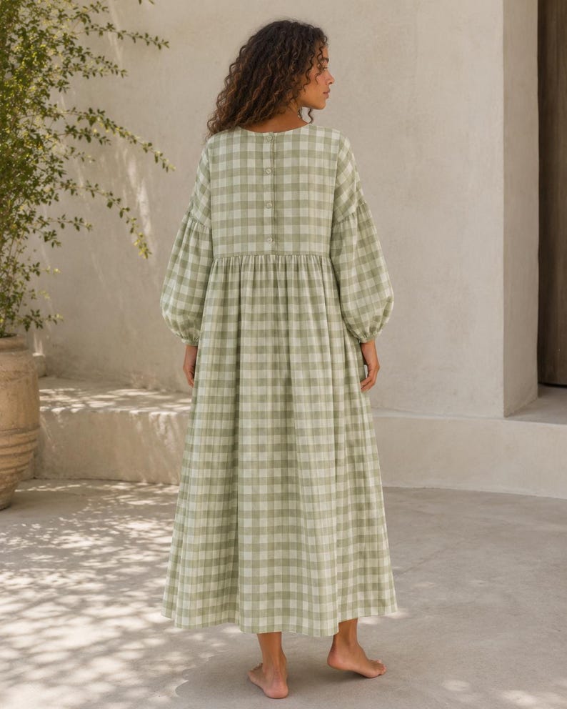 Pode incluir: Um vestido comprido at&eacute; aos p&eacute;s com um padr&atilde;o de xadrez vichy em verde s&aacute;lvia e branco. A pe&ccedil;a apresenta mangas compridas e volumosas com punhos franzidos, um decote redondo e uma fila vertical de bot&otilde;es no centro das costas.
