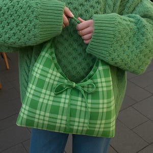 May include: A green plaid shoulder bag with a bow closure is held by a person wearing a green knit sweater and blue jeans. The bag features a light green and white plaid pattern.