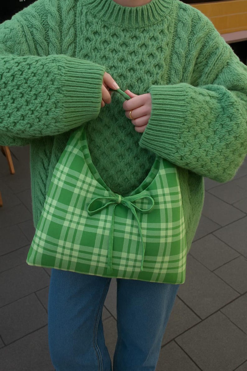 May include: A green plaid shoulder bag with a bow closure is held by a person wearing a green knit sweater and blue jeans. The bag features a light green and white plaid pattern.