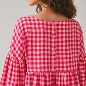 Pode incluir: Uma blusa de manga comprida com um padr&atilde;o vibrante de xadrez vichy em rosa choque e branco. A pe&ccedil;a tem um decote redondo e uma carcela vertical nas costas, presa com quatro bot&otilde;es cor-de-rosa a condizer. Inclui uma costura franzida na cintura.
