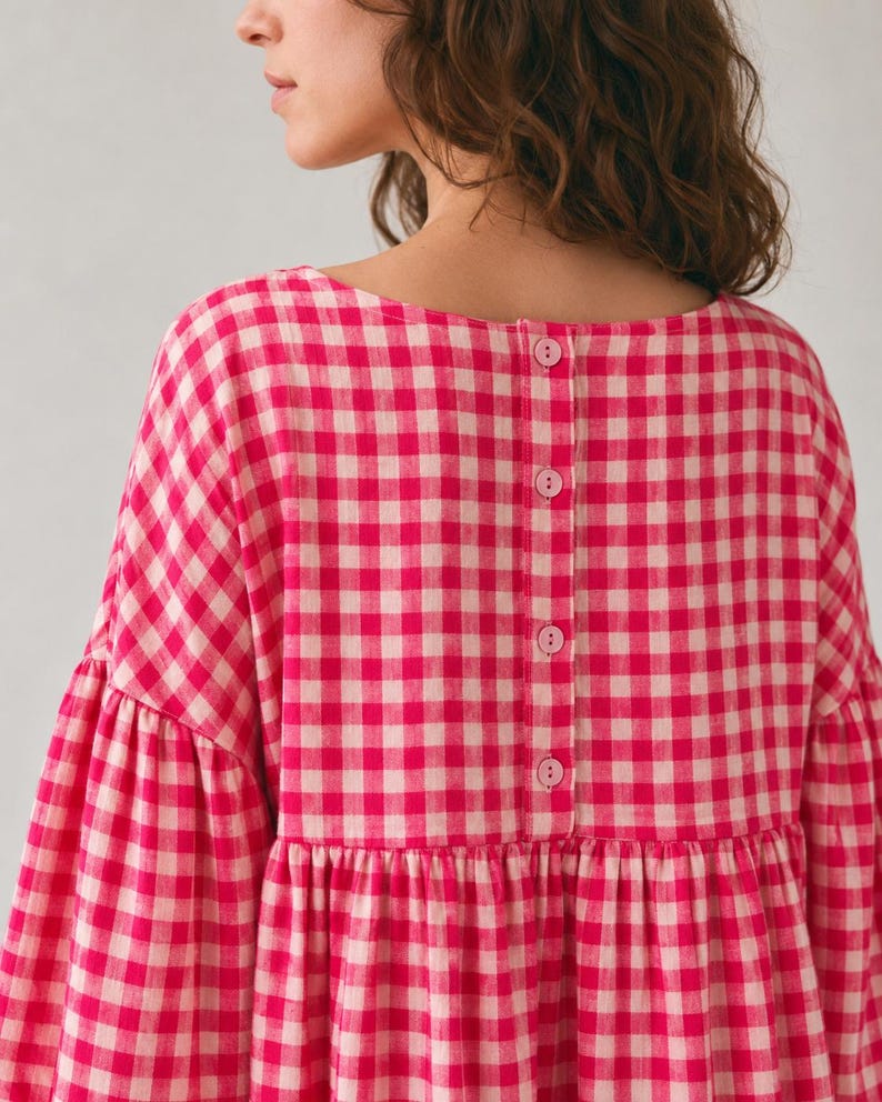 Pode incluir: Uma blusa de manga comprida com um padr&atilde;o vibrante de xadrez vichy em rosa choque e branco. A pe&ccedil;a tem um decote redondo e uma carcela vertical nas costas, presa com quatro bot&otilde;es cor-de-rosa a condizer. Inclui uma costura franzida na cintura.
