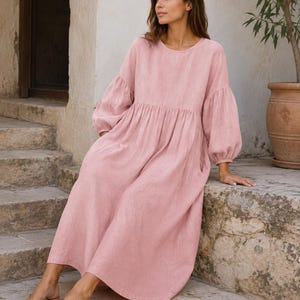 Pode incluir: Um vestido midi em rosa empoeirado com decote redondo, mangas compridas volumosas com punhos franzidos e uma silhueta relaxada e solta que cai at&eacute; ao meio da barriga da perna. A pe&ccedil;a apresenta uma costura franzida na cintura e bolsos.