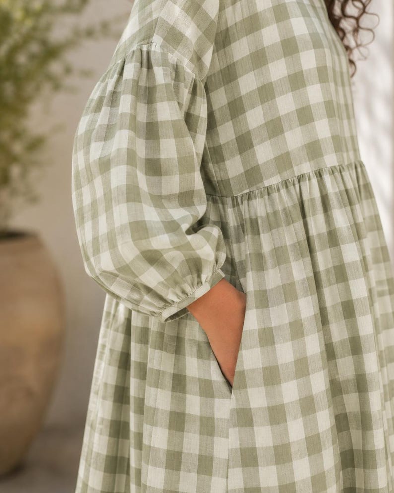 Pode incluir: Um vestido com padr&atilde;o xadrez vichy em verde s&aacute;lvia e branco, com mangas compridas e volumosas tipo bal&atilde;o com punhos franzidos e cintura franzida. O vestido inclui um bolso lateral funcional integrado na costura da saia.
