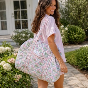 May include: A large quilted tote bag featuring a repeating floral pattern with pink flowers and green leaves on a white background, accented by a pink and white gingham check trim on the straps and the exterior pocket edge.