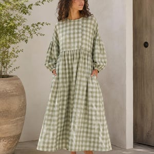 Pode incluir: Um vestido comprido at&eacute; ao ch&atilde;o de manga comprida com um padr&atilde;o de xadrez vichy em verde s&aacute;lvia e branco. A pe&ccedil;a apresenta um decote redondo, uma cintura franzida e mangas largas com punhos el&aacute;sticos, criando uma silhueta solta e fluida.