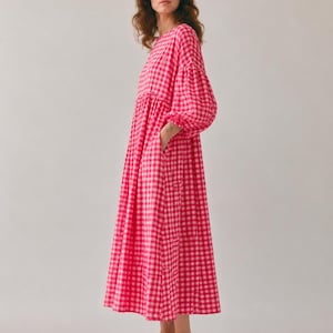 Pode incluir: Um vestido midi de manga comprida com um vibrante padr&atilde;o de xadrez vichy em rosa choque e branco. A pe&ccedil;a apresenta um decote redondo, uma cintura franzida e mangas volumosas com punhos el&aacute;sticos, caindo numa saia solta e fluida.