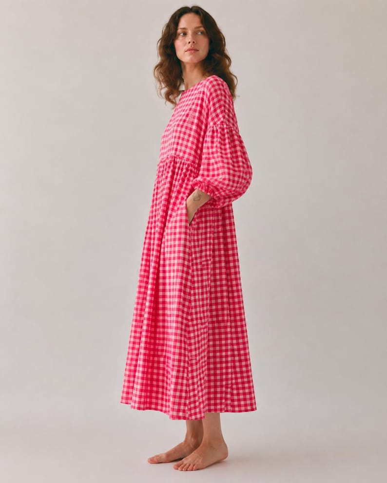 Pode incluir: Um vestido midi de manga comprida com um vibrante padr&atilde;o de xadrez vichy em rosa choque e branco. A pe&ccedil;a apresenta um decote redondo, uma cintura franzida e mangas volumosas com punhos el&aacute;sticos, caindo numa saia solta e fluida.