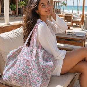May include: A quilted tote bag featuring a repeating floral pattern in shades of pink, green, and blue on a white background. The bag has two shoulder straps with a pink and white gingham check trim that matches the piping along the seams.