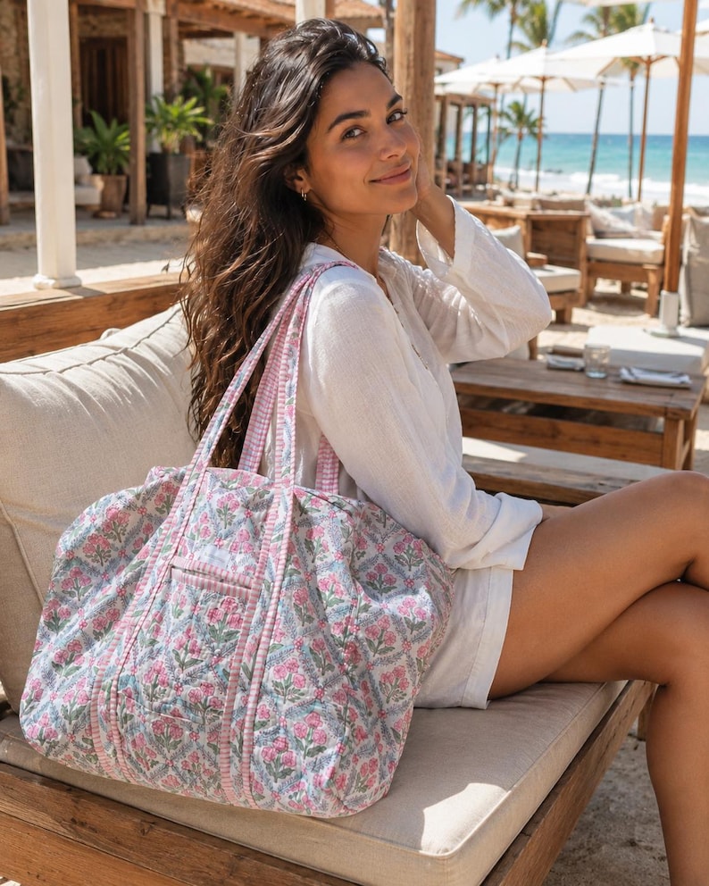 May include: A quilted tote bag featuring a repeating floral pattern in shades of pink, green, and blue on a white background. The bag has two shoulder straps with a pink and white gingham check trim that matches the piping along the seams.