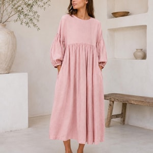Pode incluir: Um vestido midi em rosa empoeirado com decote redondo, mangas compridas volumosas com punhos franzidos e uma cintura relaxada e franzida que flui para uma saia larga e solta com bolsos laterais, feito de um tecido texturizado.