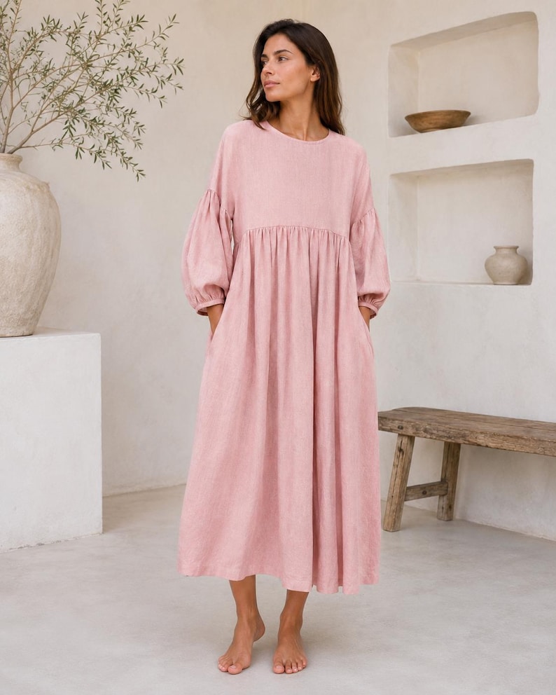 Pode incluir: Um vestido midi em rosa empoeirado com decote redondo, mangas compridas volumosas com punhos franzidos e uma cintura relaxada e franzida que flui para uma saia larga e solta com bolsos laterais, feito de um tecido texturizado.