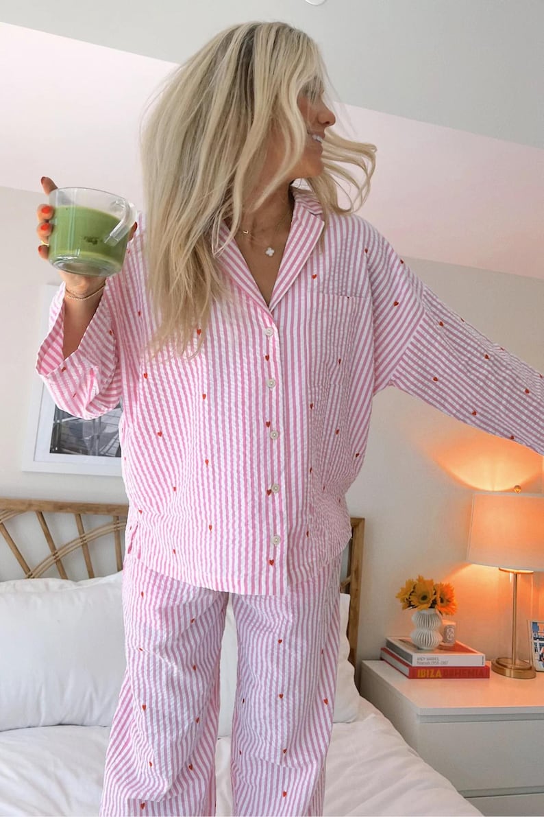 May include: A two-piece pajama set featuring a long-sleeved button-down shirt and matching pants. The fabric is white with vertical pink stripes and small, scattered red heart embroidery throughout the entire garment, including a chest pocket.