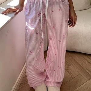 May include: Light pink and white striped lounge pants featuring a repeating pattern of small pink bows. The pants have a textured seersucker-style fabric, a wide elastic waistband with a white drawstring tie, and a loose, relaxed leg fit.